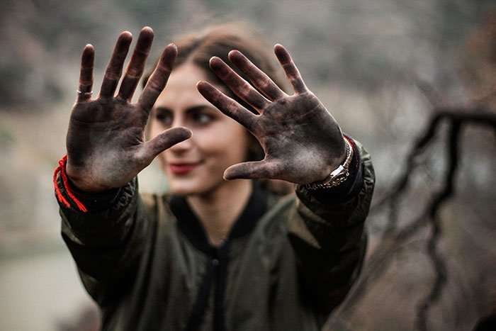 A person with dark hair is slightly out of focus in the background, holding up their hands in front of their face. Their palms are covered in dirt, and they are wearing a green jacket and red bracelets. The scene looks like an outdoor, natural setting, reminiscent of a quality website developer's creative spirit.